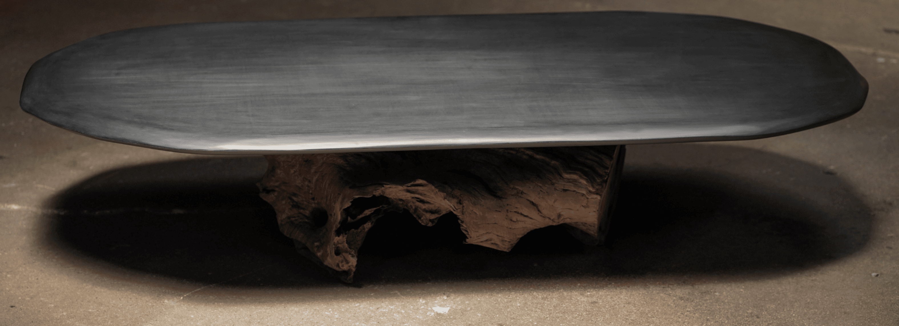 A low coffee table featuring an elongated oval top made of dark, matte material, possibly stone, supported by a unique and irregular base crafted from a natural wood root or stump. The design combines a sleek modern top with an organic, sculptural base. — 3D model
