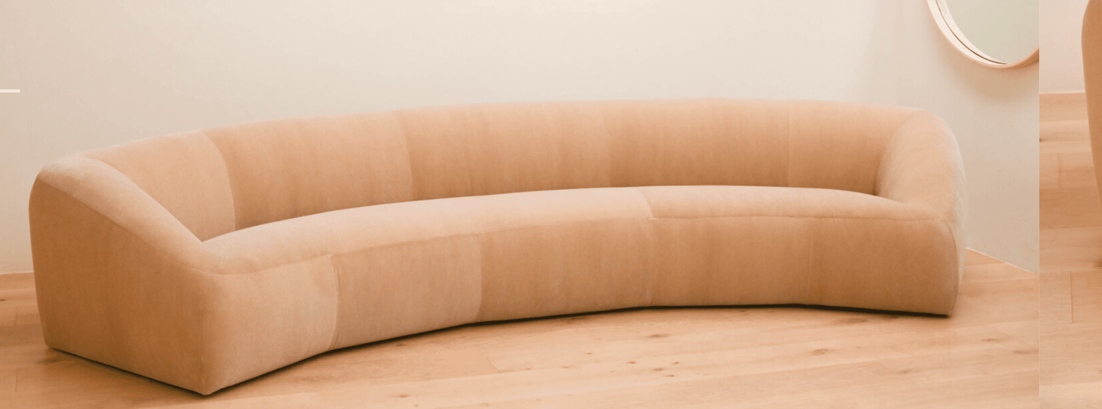 A contemporary, generously curved upholstered sofa in a light, neutral beige tone. The sofa features a continuous, rounded form with no visible legs, giving it a low-profile and substantial presence. The backrest and seat are seamlessly integrated, suggesting a cohesive, single-piece construction. The upholstery appears to be a textured fabric, possibly boucle or a fine-grained velvet, with subtle vertical seams segmenting the back and seat portions. Its design is minimalist, focusing on organic curves and comfort. — 3D model