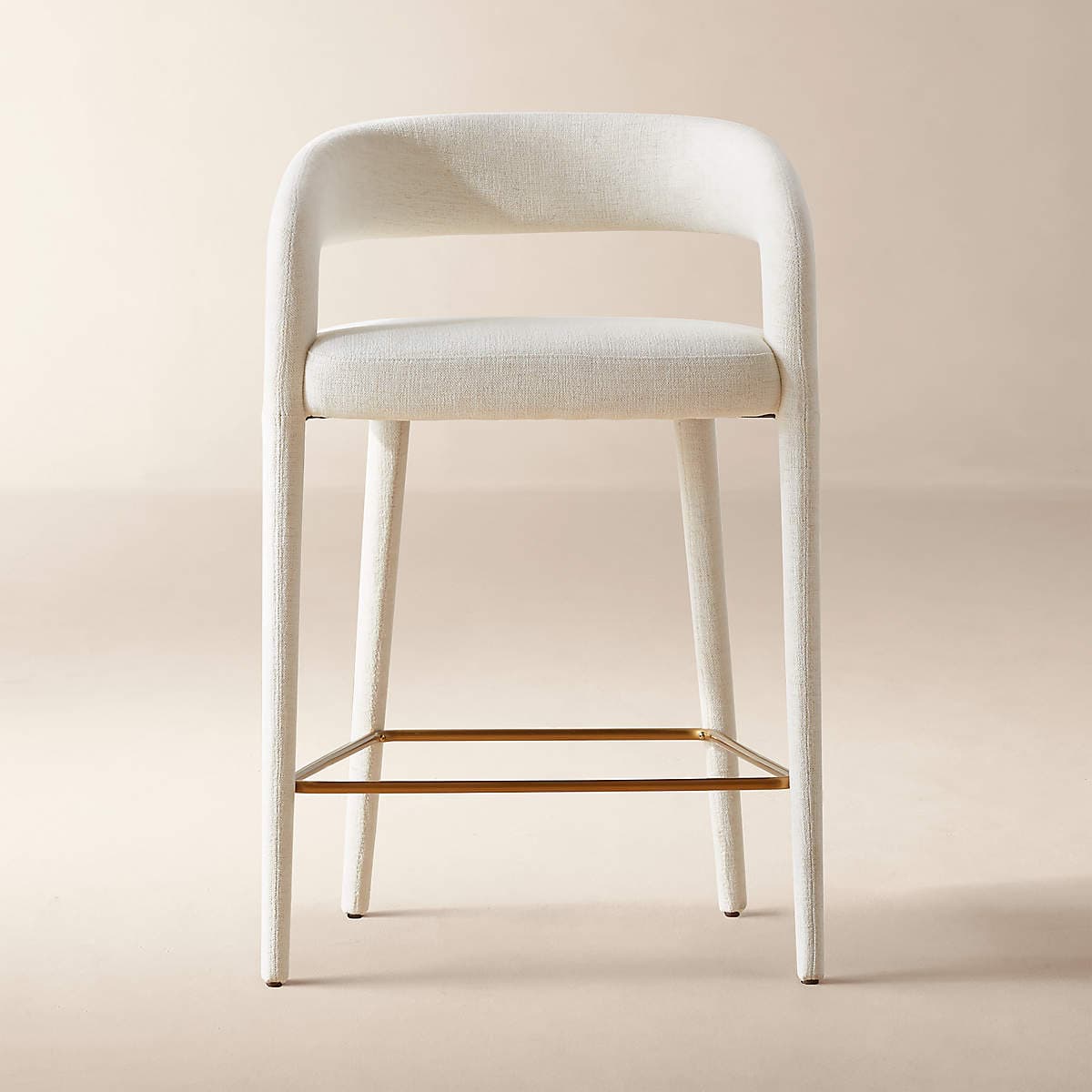 A contemporary bar stool featuring a fully upholstered body in an off-white textured fabric. The stool has a curved, open backrest that extends into the seat. Its four straight legs are also fully upholstered and are connected by a square brass-toned metal footrest, providing stability and a contrasting accent. — 3D model