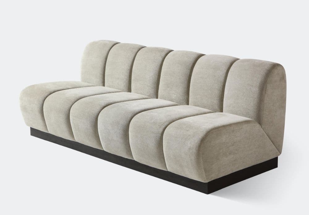 A contemporary armless sofa featuring distinct channel-tufted upholstery that forms rounded, individual sections across both the seat and backrest. The sofa is finished in a light, neutral-toned, velvet-like fabric and rests upon a recessed, dark plinth base. — 3D model