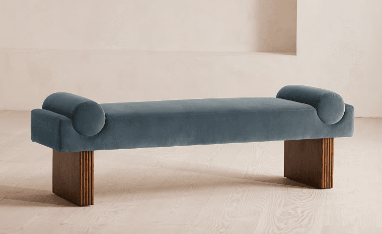 A modern daybed featuring a long, rectangular upholstered cushion with cylindrical bolsters integrated into each end. The seating surface is covered in a plush teal/slate blue velvet fabric. It rests upon two substantial, rectangular block legs made of dark-toned wood, characterized by a fluted or reeded texture on their outward-facing sides. The design is backless, emphasizing a sleek, contemporary profile. — 3D model
