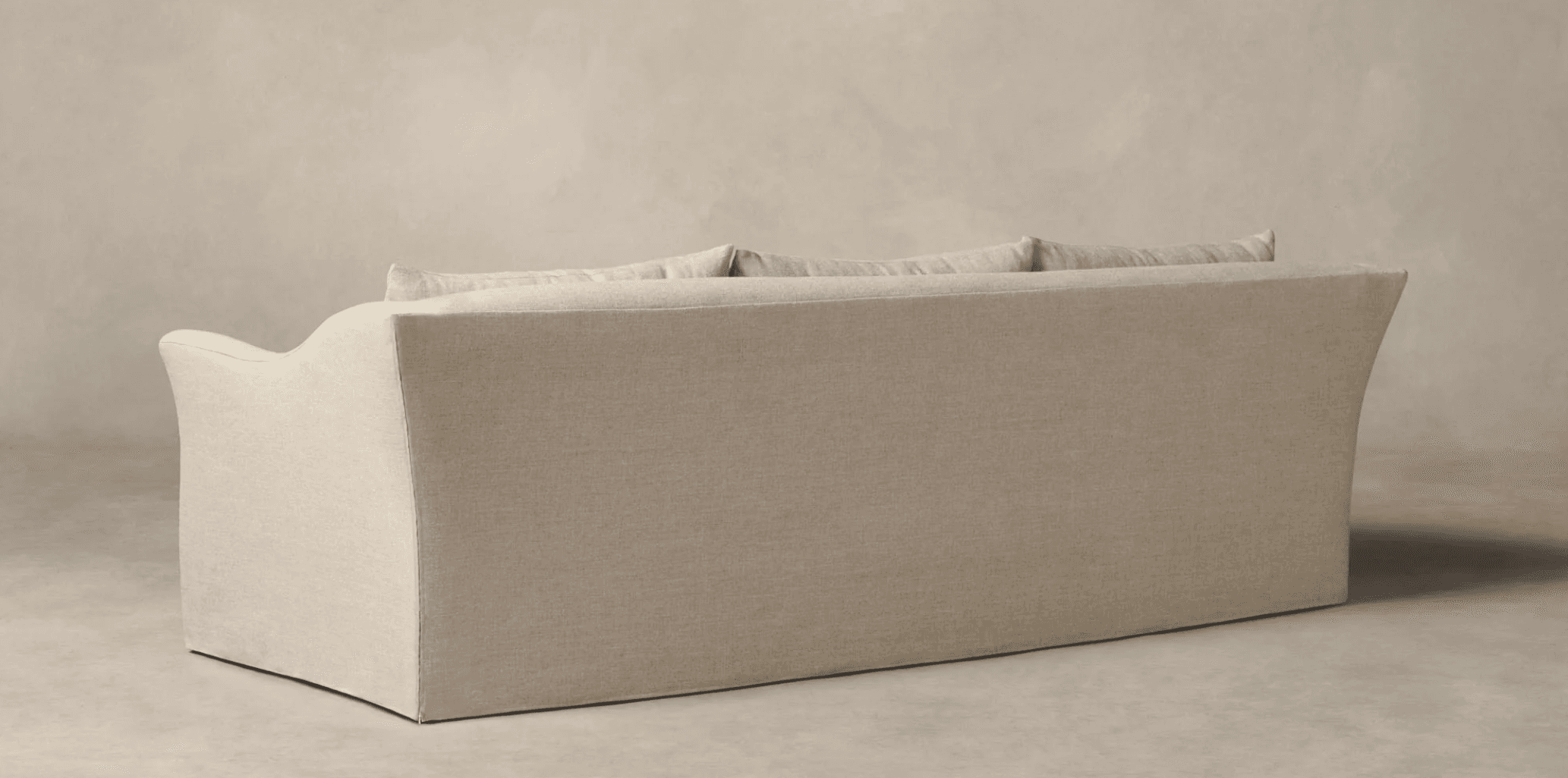 A three-seater sofa, viewed from the rear and side. The sofa features a loose slipcover construction with a skirted base that extends to the floor. It has a low profile with a gently curved armrest visible on the left. The back is composed of multiple cushions, suggesting a comfortable and plush design. The upholstery appears to be a textured linen or linen-blend fabric. — 3D model