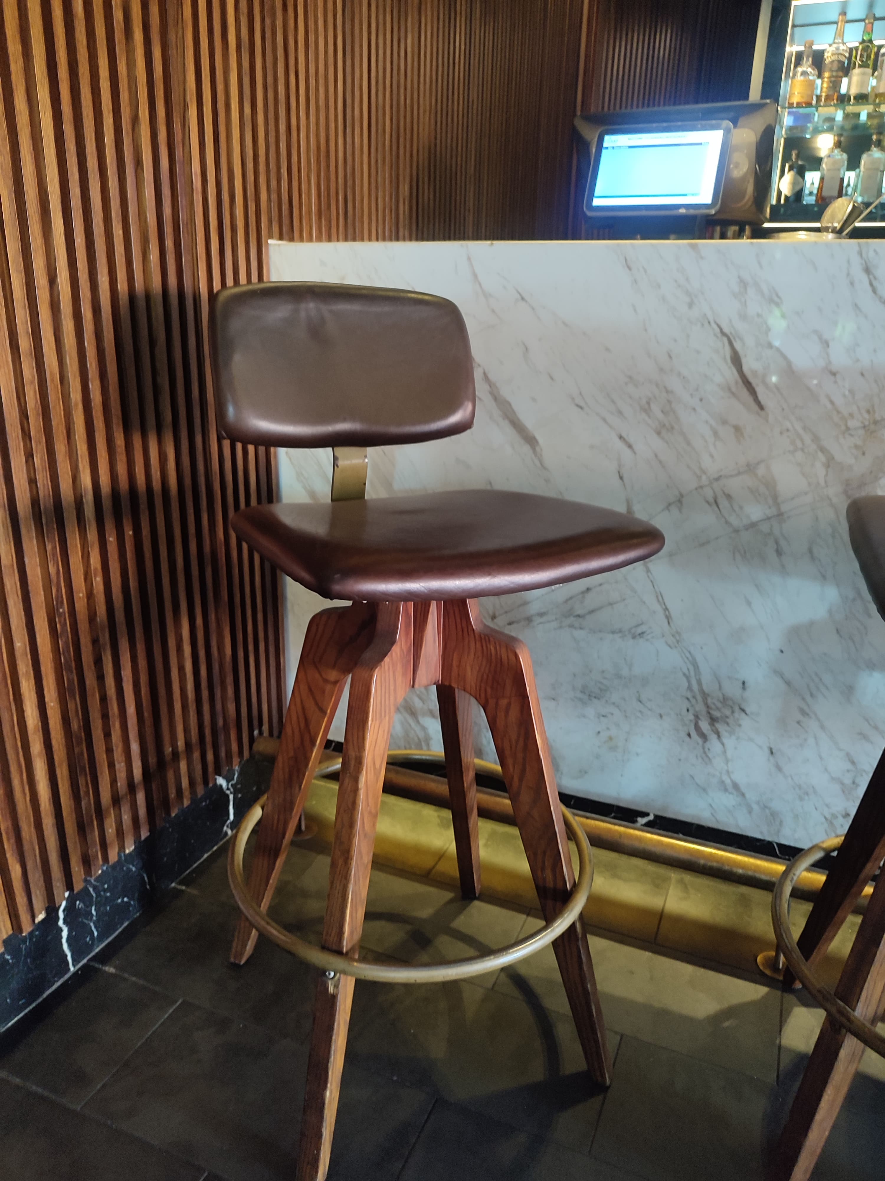 A brown bar stool with a swiveling seat and backrest, supported by a wooden tripod base. The seat and backrest appear to be upholstered. A brass-colored metal ring encircles the base of the legs, serving as a footrest. The overall design suggests a mid-century modern aesthetic. — 3D model