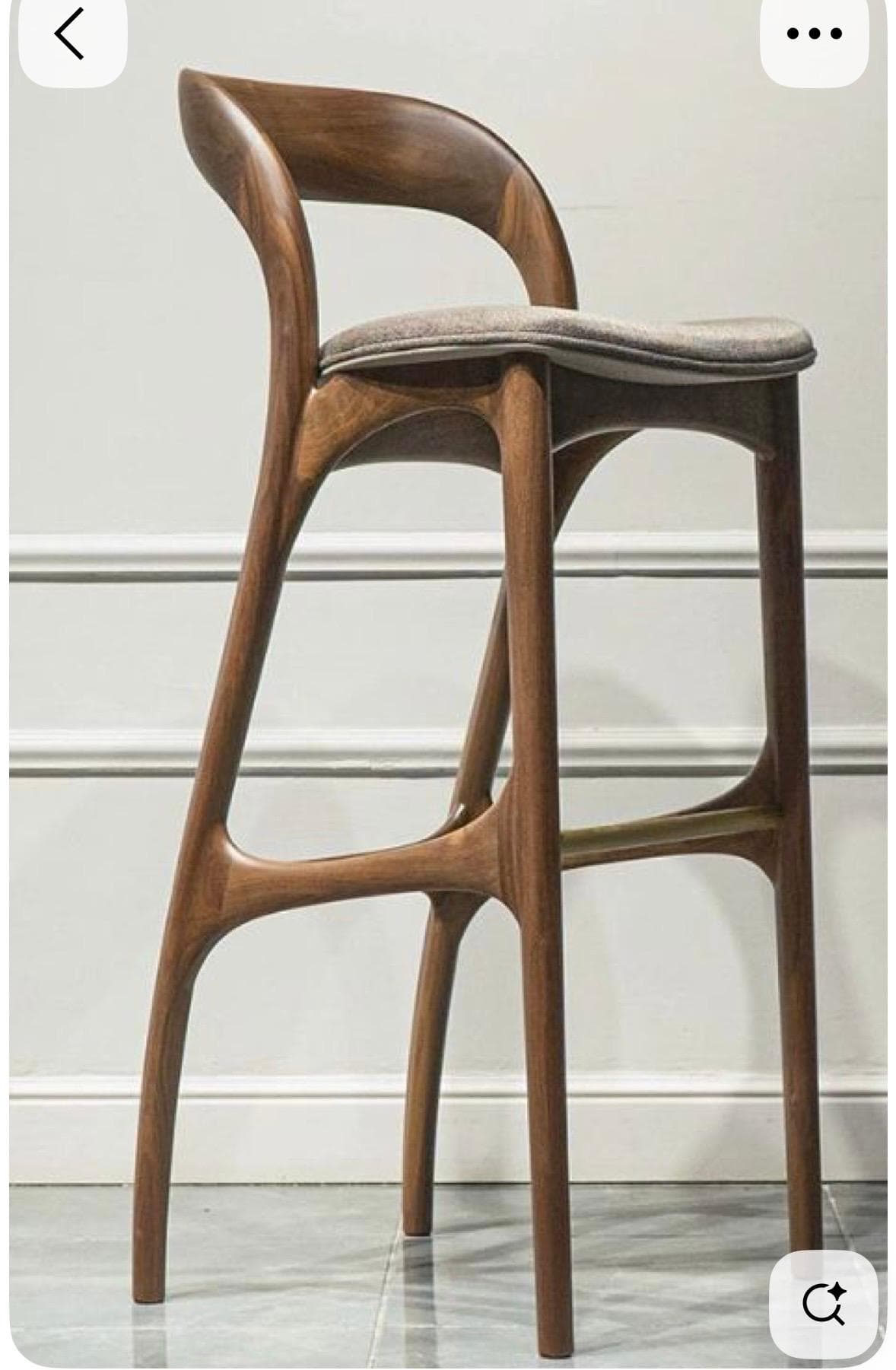 A contemporary bar stool with a dark walnut wood frame, featuring gracefully curved elements forming the backrest and legs. It has a padded, light grey fabric upholstered seat and a brass-finished metal footrest. — 3D model