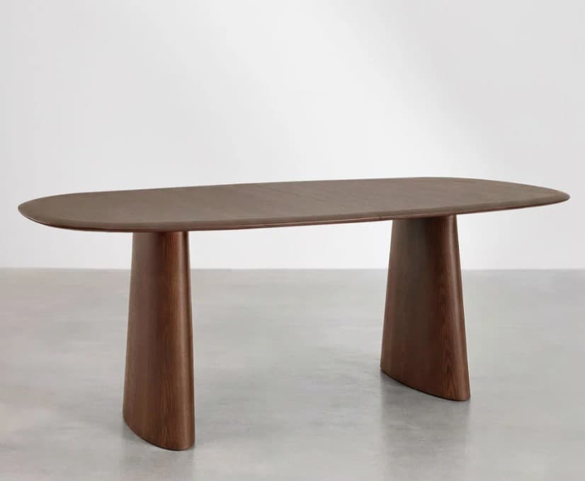 A modern dining table with an elongated, oval-shaped tabletop supported by two distinct, sculptural pedestal bases. The table exhibits a contemporary design with clean lines and a minimalist aesthetic. The material appears to be wood with a dark stain, showcasing a visible wood grain pattern. — 3D model