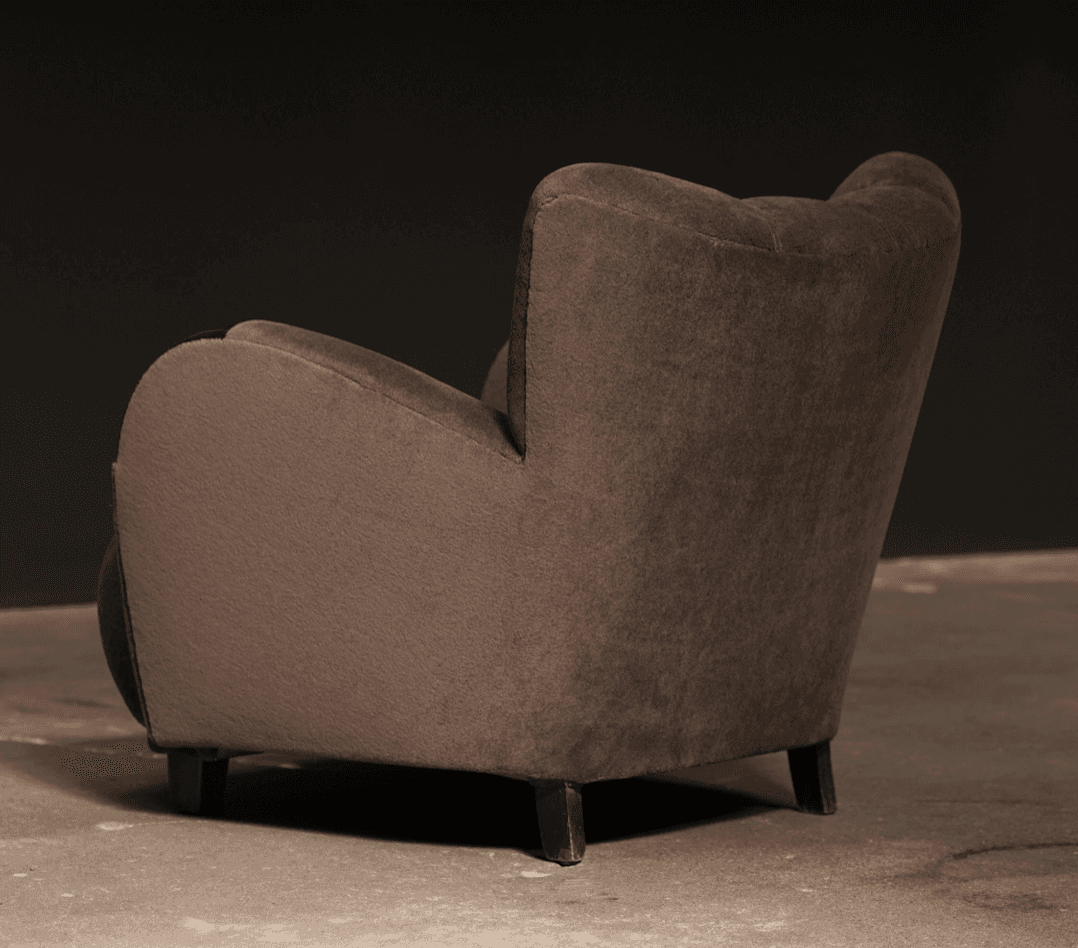 A robust, fully upholstered armchair viewed from the rear-right side, featuring a rounded backrest, integrated arms, and short, dark wooden legs. The chair is covered in a soft, dark taupe fabric. — 3D model