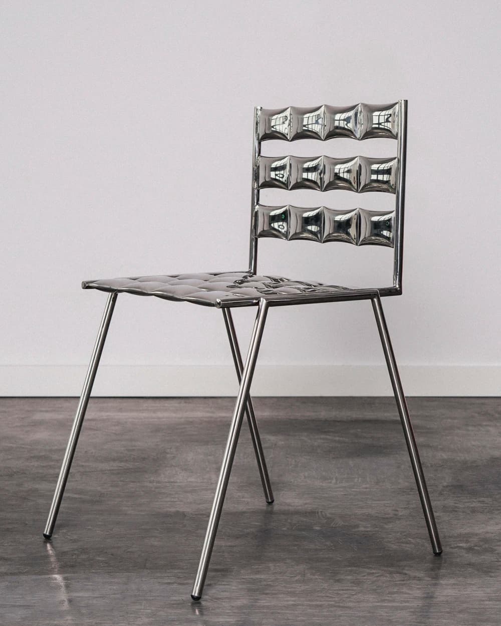 A modern chair with a distinctive design featuring a highly reflective, metallic finish. The seat and backrest are composed of multiple square-shaped, dimpled elements arranged in a grid pattern. The frame and four slender legs are also metallic, providing a sleek and contemporary aesthetic. The legs are angled outwards for stability. — 3D model