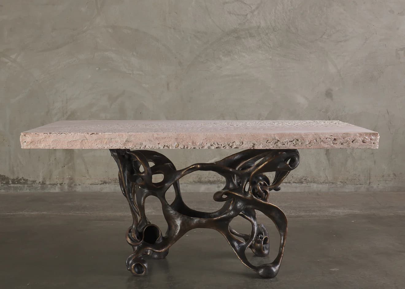 A console table with a rectangular, light pinkish-beige stone top and an intricately sculpted, dark bronze base. The base features an organic, abstract design with interconnected hollow forms and flowing lines, resembling bones or skeletal structures. The stone top has a rough-hewn edge and a textured surface with visible inclusions. — 3D model