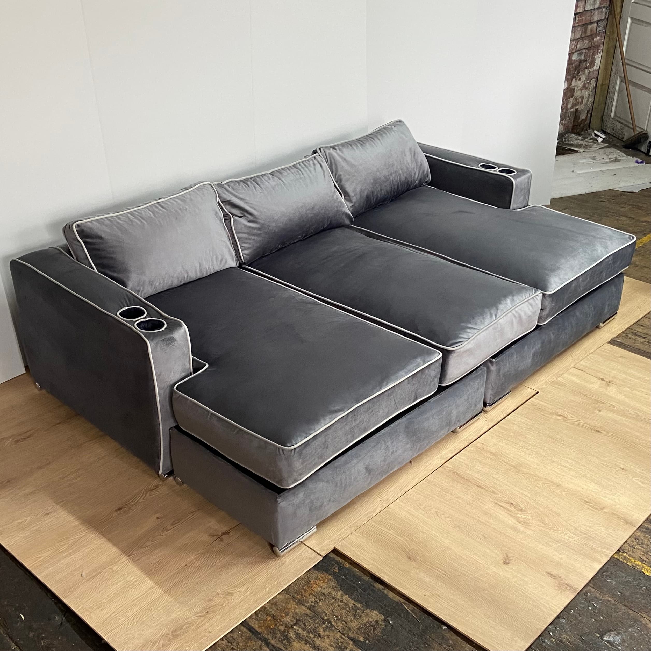 A modern sectional sofa with a chaise lounge extension on one side. The sofa features multiple seat cushions and back cushions, upholstered in a soft, plush gray fabric. It has a piped trim in a lighter contrasting color along the edges of the cushions and frame. Two integrated cup holders are visible on the armrests. The base appears to be a solid structure, possibly with decorative metal feet. — 3D model