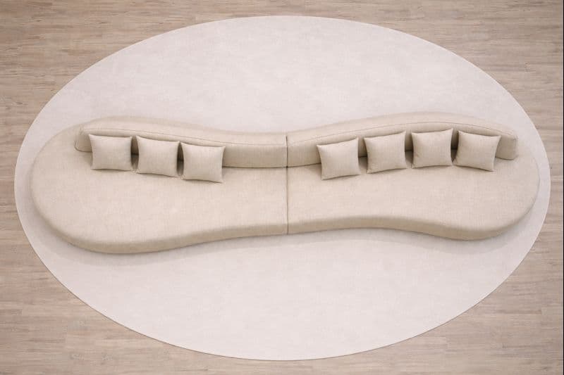 A large, modular, curvilinear sofa with a neutral-toned fabric upholstery. The sofa is composed of multiple sections that form an S-shape or wave-like contour. It features integrated armrests and back support along its length. Seven square throw pillows with matching upholstery are strategically placed along the back of the sofa. The sofa appears to be situated on a large, light-colored circular rug. — 3D model