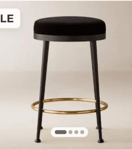 A modern bar stool featuring a round, thickly upholstered black velvet or fabric seat. It stands on four slender, tapered black metal legs, reinforced by a circular brass-finished metal footrest. The design is minimalist and contemporary. — 3D model