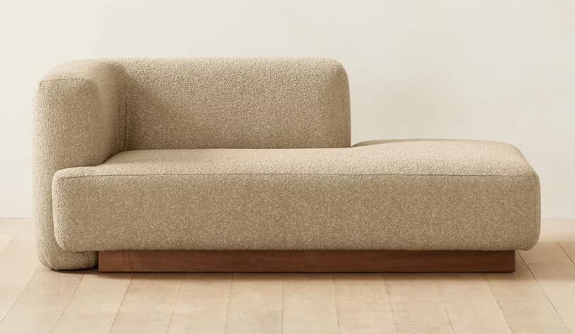 A modern upholstered chaise lounge featuring a single integrated left armrest and a low-profile design. The piece is covered in a textured light beige fabric and rests on a recessed wooden plinth base. — 3D model