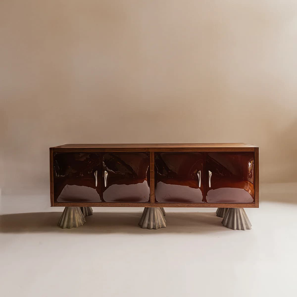 A long, rectangular sideboard featuring a walnut wood frame and top surface. The front is composed of four individual cabinet doors with a high-gloss, deep reddish-brown lacquered finish. Each pair of doors is equipped with two horizontal, silver-toned bar pulls. The entire cabinet is elevated on four distinctive, sculptural, conical legs with a fluted or pleated texture, appearing to be made from a light-colored composite or concrete material. The design combines classic wood elements with contemporary finishes and an abstract base. — 3D model