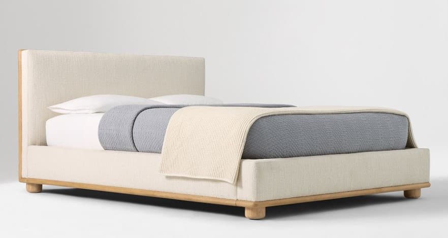 A contemporary bed frame with a fabric-upholstered headboard and base, featuring a visible wooden frame accent around the perimeter and cylindrical wooden legs. The bed is dressed with white bedding, a grey textured blanket, and a cream-colored knitted throw blanket. — 3D model