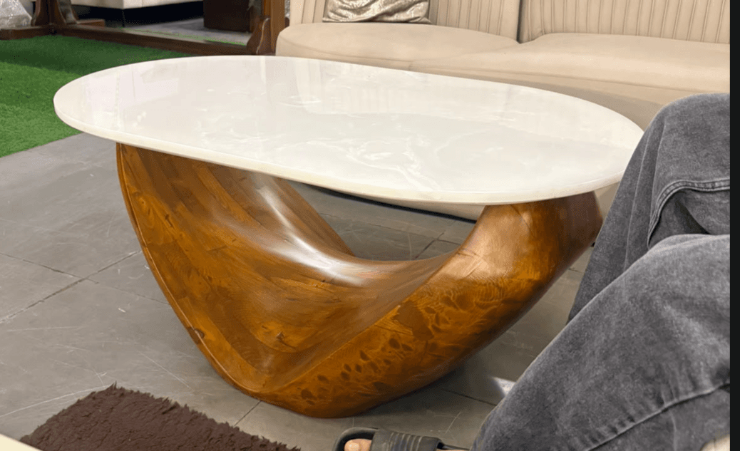 An organic modern coffee table featuring a large, smooth oval white tabletop resting on a dramatically sculpted and continuous wooden base. The base is crafted from warm-toned wood, showcasing natural grain patterns and a flowing, abstract design that appears to cradle the top. — 3D model