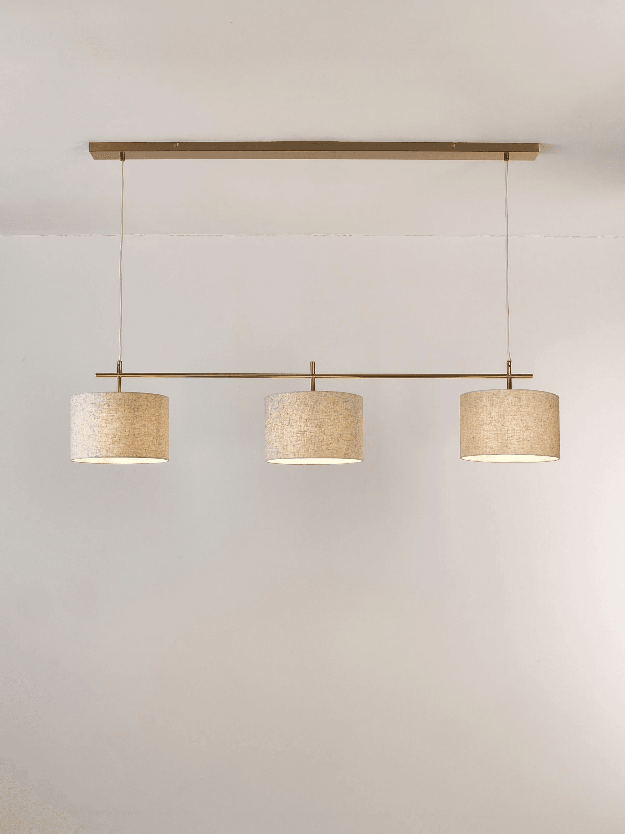 A linear pendant light fixture with a rectangular brass-finished metal bar mounted horizontally to the ceiling. Three cylindrical fabric shades are suspended from the bar by thin metal rods and white electrical cords. Each shade has a textured, linen-like appearance and emits a warm light. — 3D model
