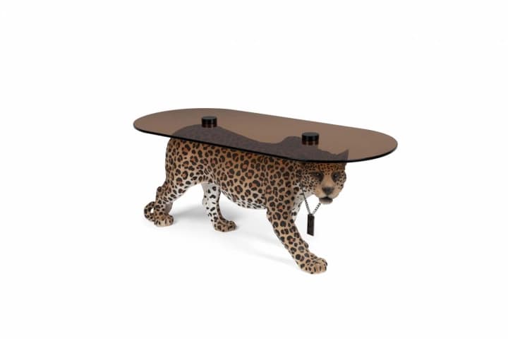 An artistic coffee table designed in the shape of a walking leopard. The leopard's body serves as the base, supporting an oval-shaped, smoked glass tabletop. The leopard figure is detailed with a realistic spotted pattern. A decorative chain with a pendant hangs from the leopard's neck. — 3D model