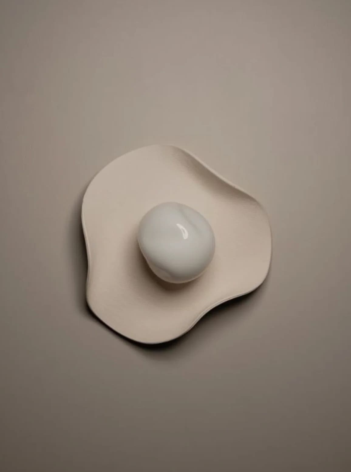 A modern wall sconce featuring an irregular, organic-shaped backplate in a matte cream or beige finish, with a central spherical diffuser made of white, translucent material, possibly glass or marble. The design evokes a minimalist and sculptural aesthetic. — 3D model