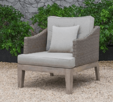 A contemporary outdoor armchair featuring a sturdy wooden frame and tightly woven synthetic rattan armrests and back. The chair is equipped with thick, plush seat and back cushions, along with an additional accent pillow, all upholstered in a light greige fabric. The wooden frame has a light, natural finish, complementing the greyish-brown tone of the woven elements. The overall design emphasizes comfort and durability, suitable for outdoor living spaces. — 3D model