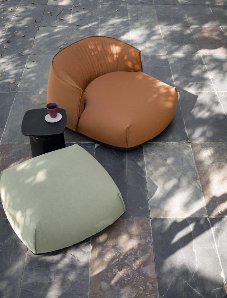 A modern armchair with a rounded, low-profile design. The seat and back are upholstered in a soft, textured material, with the back featuring subtle pleating. It has a low, dark-colored base that appears to be made of wood or a similar composite material. — 3D model
