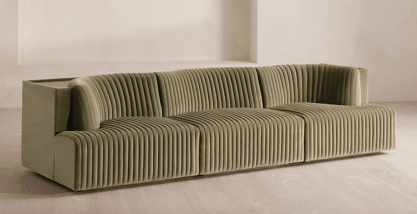 A long, contemporary sofa with a channeled upholstery design. The sofa features a modular appearance with distinct sections, creating a sense of depth and texture. It has a low profile and a deep seat, with curved armrests or side panels. A small, dark wood-toned surface is visible on the far left side, integrated into the structure. — 3D model