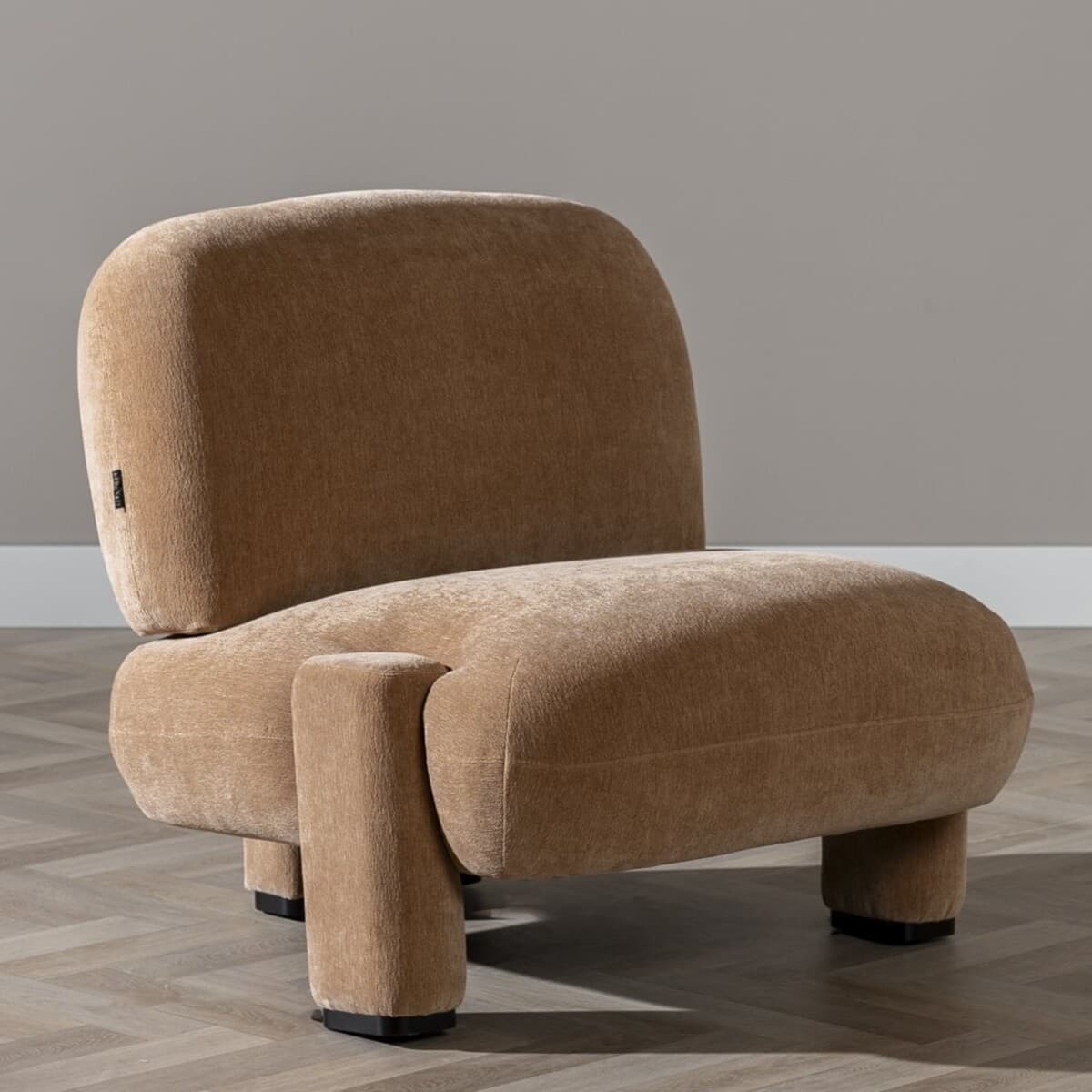 A contemporary armchair with a rounded, oversized silhouette. The chair features a low profile with generously padded upholstery and integrated armrests that merge into the main body. It is supported by four block-like legs, two of which are clearly visible and extend from the sides of the seat base. A small black tag is attached to the side of the backrest. — 3D model