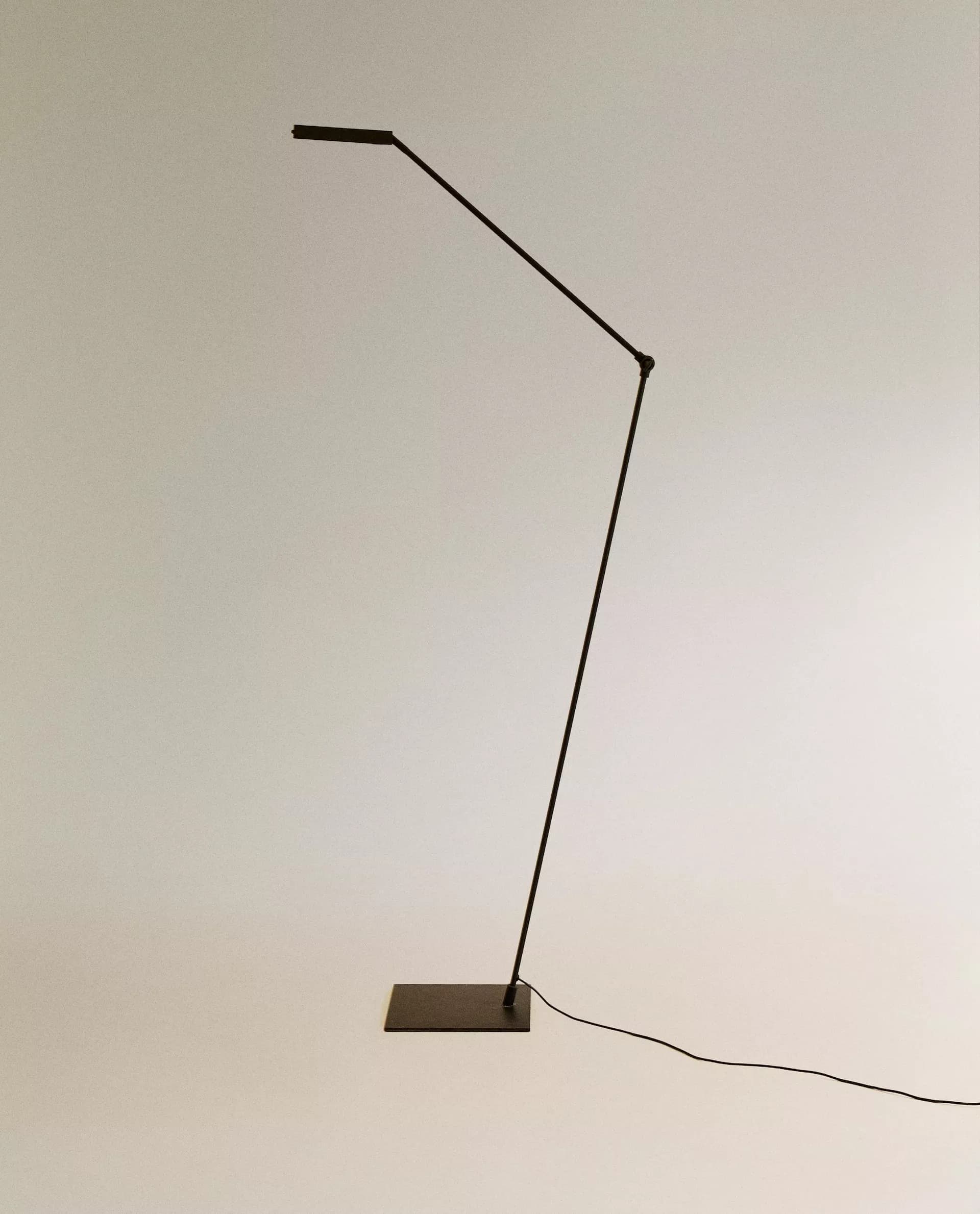 A modern and minimalist floor lamp featuring a slender, dark-finished metal body with an adjustable arm and a flat, rectangular base. The light source is an integrated LED panel, covered by a translucent diffuser, providing a clean and functional aesthetic. A power cord extends from the base. — 3D model