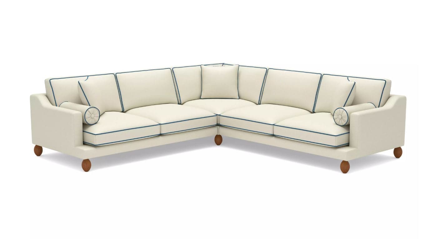 A sectional sofa with a corner configuration. It features a light-colored upholstery with contrasting dark blue piping along the edges of the cushions and frame. The sofa includes several back cushions and armrest cushions, with one cylindrical bolster pillow on the left armrest. It is supported by four short, rounded wooden legs. — 3D model