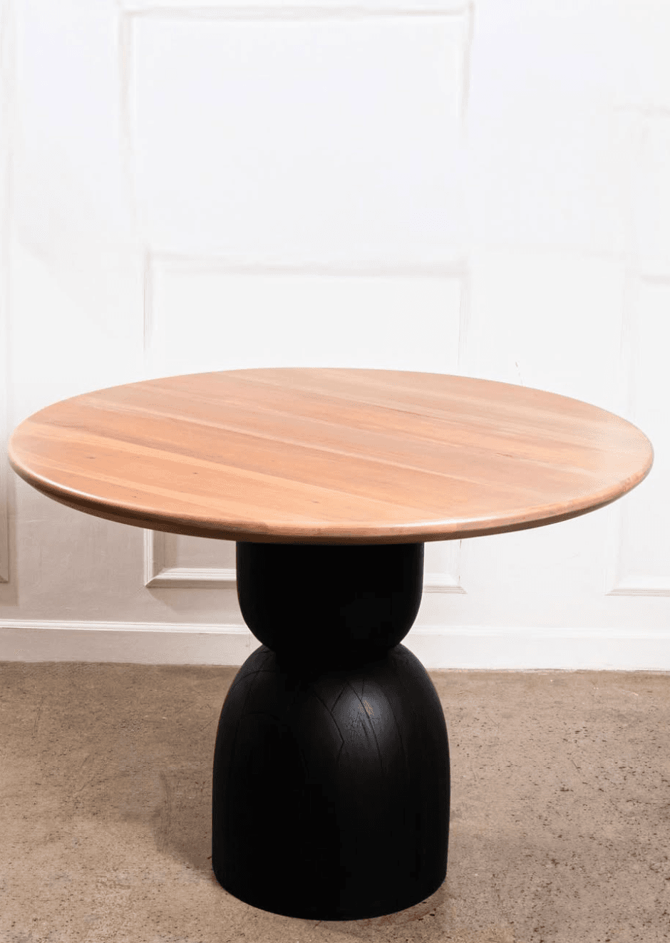 A contemporary round side table featuring a light-toned wooden top and a matte black sculptural pedestal base. The base has a distinctive hourglass-like shape with subtle vertical lines or grooves, giving it a tactile texture and modern aesthetic. The table presents a two-tone appearance, blending natural wood with a bold black finish. — 3D model