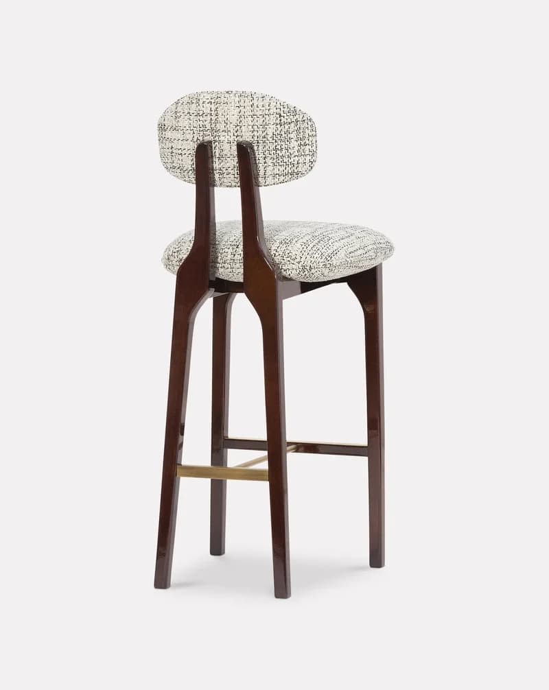 A high-backed bar stool featuring a dark stained wooden frame, a contoured upholstered seat, and a separate, floating upholstered backrest. The frame includes splayed legs and a brass footrest. The upholstery is a textured, patterned fabric in monochrome tones. — 3D model