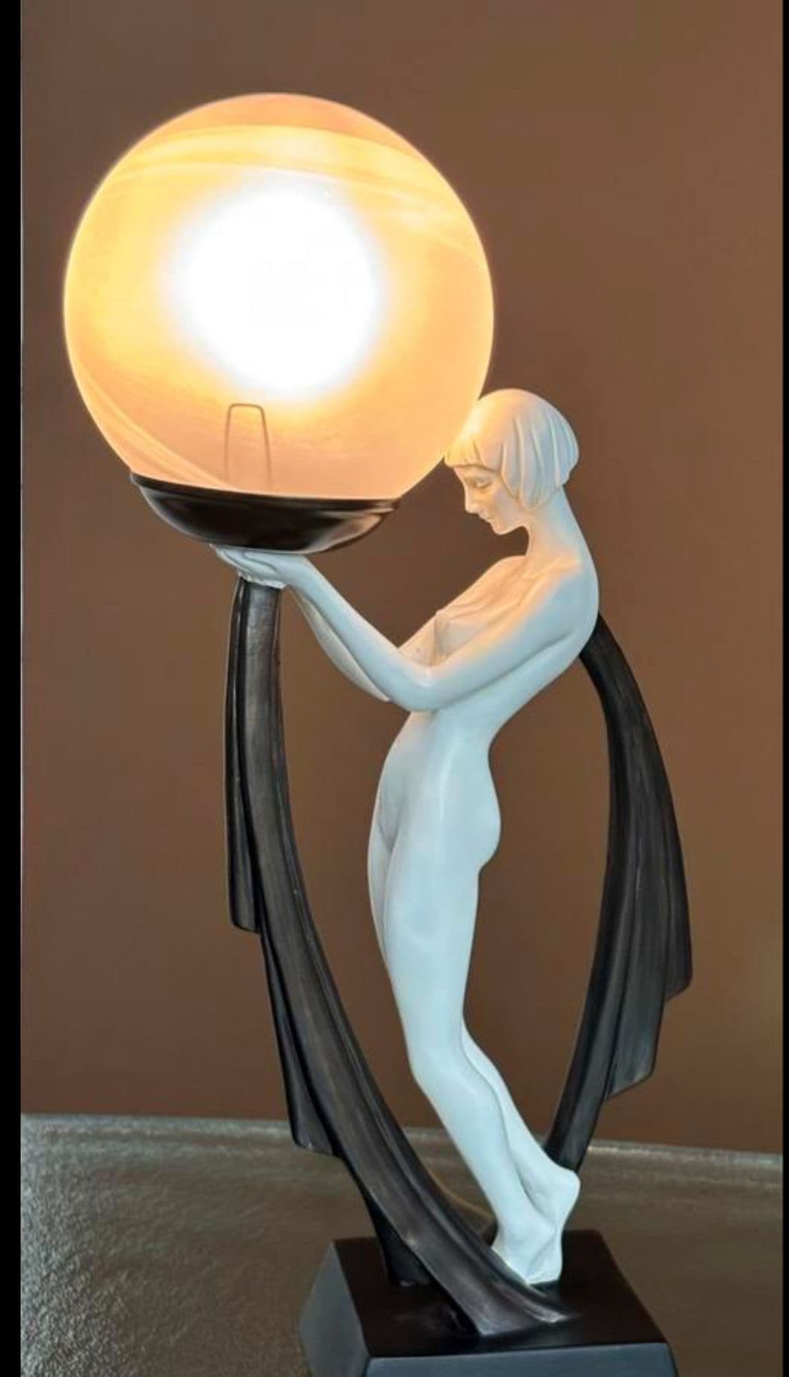 A striking Art Deco style figural table lamp featuring a nude female figure in a light finish, gracefully supporting a large, spherical frosted glass shade. The figure is integrated into a dark, flowing base and support structure with geometric accents, creating a high-contrast decorative lighting piece. — 3D model