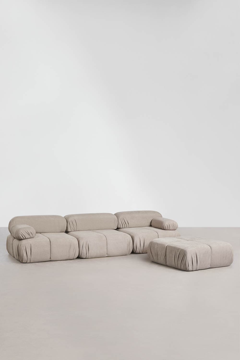 A light beige, plush modular sofa with an accompanying ottoman, featuring a contemporary design with soft, rounded forms and distinctive gathered fabric detailing around the base of each section. The sofa is composed of multiple configurable units, creating a flexible seating arrangement. — 3D model