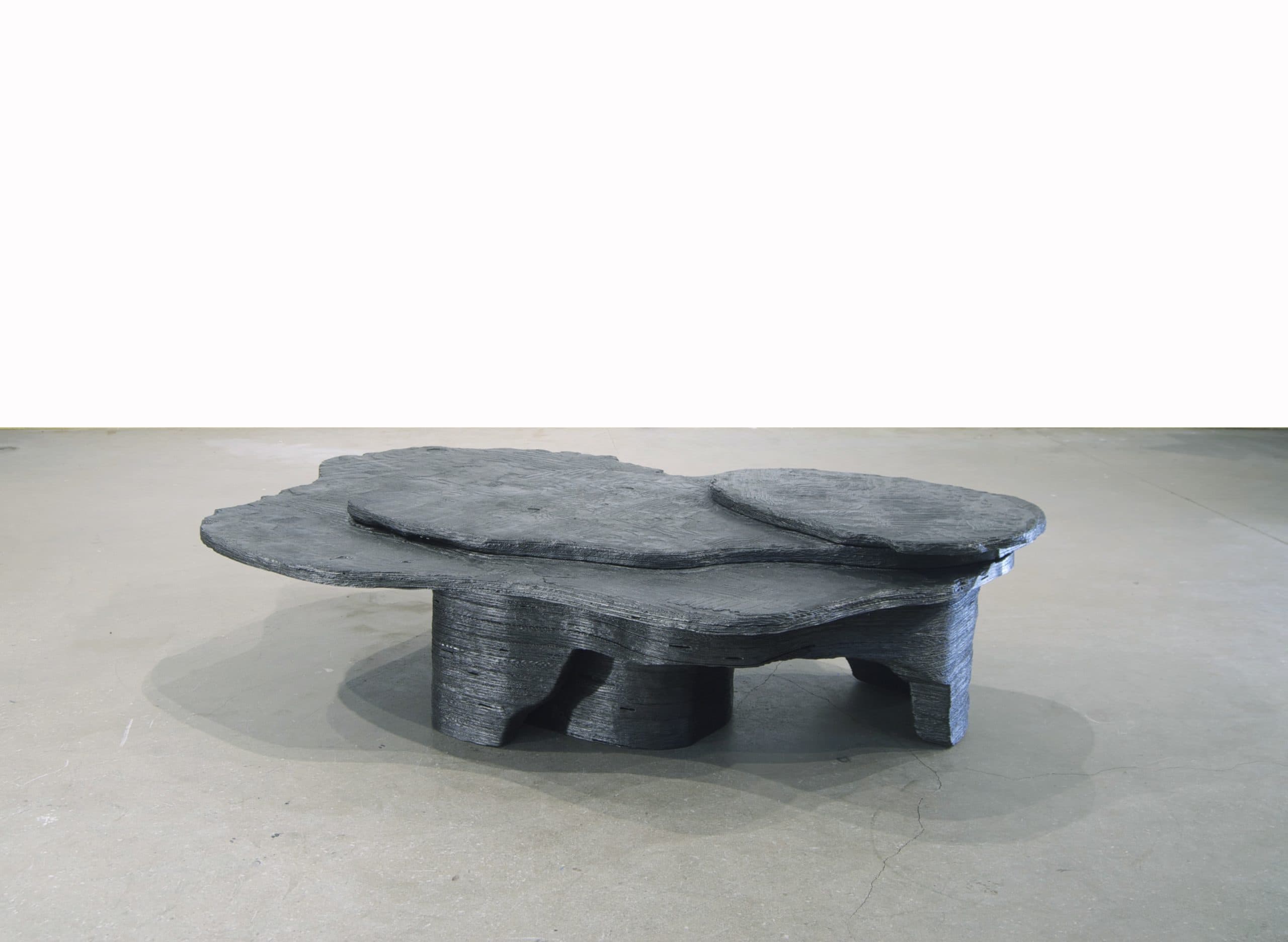 A sculptural, low-lying table featuring an organic, multi-tiered design. The dark, matte material has a layered texture, mimicking natural rock formations or topographical maps. The table top comprises several irregularly shaped, overlapping planes at varying heights, supported by an equally irregular and textured base. — 3D model