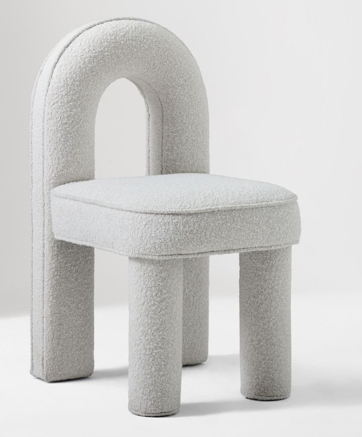 A contemporary chair featuring a distinctive arched backrest and a square seat, all fully upholstered in a light gray or off-white boucle fabric. The chair is supported by four robust, tubular legs, also fully upholstered, giving it a soft, sculptural, and monolithic appearance. The design emphasizes rounded forms and a plush texture. — 3D model