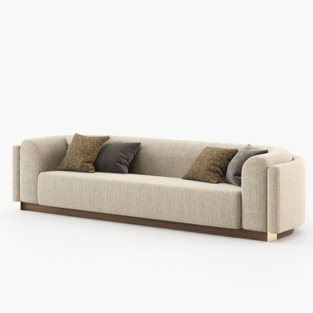 A contemporary three-seater sofa with a continuous back and armrest design, upholstered in a light beige textured fabric. It features rounded armrests, a prominent dark wood plinth base, and subtle brass accents on the lower outer edges of the arms. Several toss pillows in brown and gray are included. — 3D model