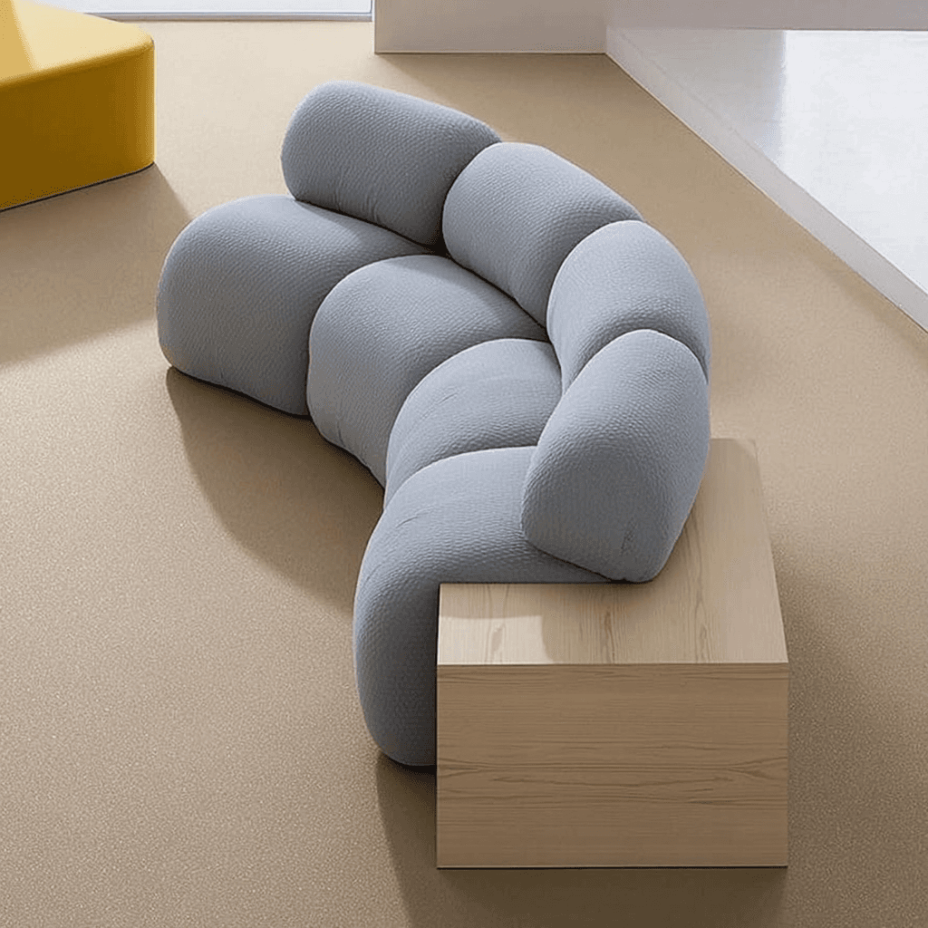 A modular sectional sofa with a curved design. The seating elements are rounded and puffy, resembling stacked cushions or pillows. It is upholstered in a light grey, textured fabric. Attached to one end is a rectangular wooden module that serves as an end table or armrest extension. The wood appears to be light-colored with a visible grain pattern. — 3D model
