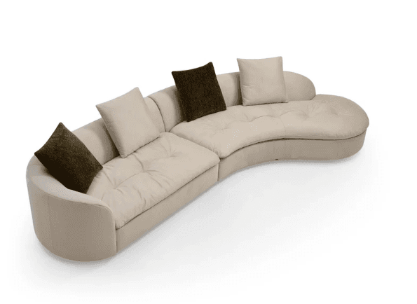 A contemporary curved sectional sofa, upholstered in a light beige or cream fabric. The sofa features a low-profile backrest that curves smoothly along its length, creating a crescent shape. The seat cushions are integrated and exhibit subtle button tufting, providing a tailored yet plush appearance. The design appears modular, with distinct sections forming the overall curve. Four throw pillows, two light beige and two dark brown, are placed on the sofa, complementing its neutral palette. The base of the sofa is concealed, suggesting a floor-level design, and the overall aesthetic is sleek and modern. — 3D model