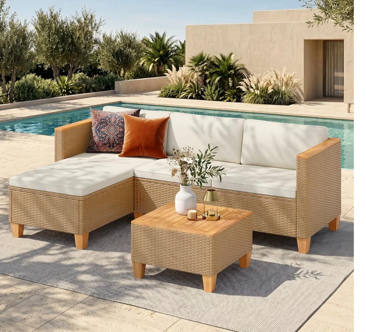 A modern outdoor L-shape sectional sofa set featuring a chaise lounge section, a three-seater section, and a matching low coffee table. The frames of the sofa and table are constructed from woven light brown synthetic rattan or natural wicker, complemented by light wood armrests, table top slats, and block legs. The sofa is outfitted with thick, light cream-colored seat and back cushions. Two decorative throw pillows, one patterned and one solid orange, are placed on the chaise lounge. The set is designed for outdoor use, offering comfortable and stylish seating. — 3D model