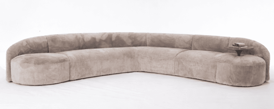 A large, curved sectional sofa with a smooth, plush upholstery. The sofa features a rounded, low-profile design with integrated armrests and a continuous seating surface that forms a semi-circle. A small, circular side table with a dark finish and two books is placed at one end of the sofa. — 3D model