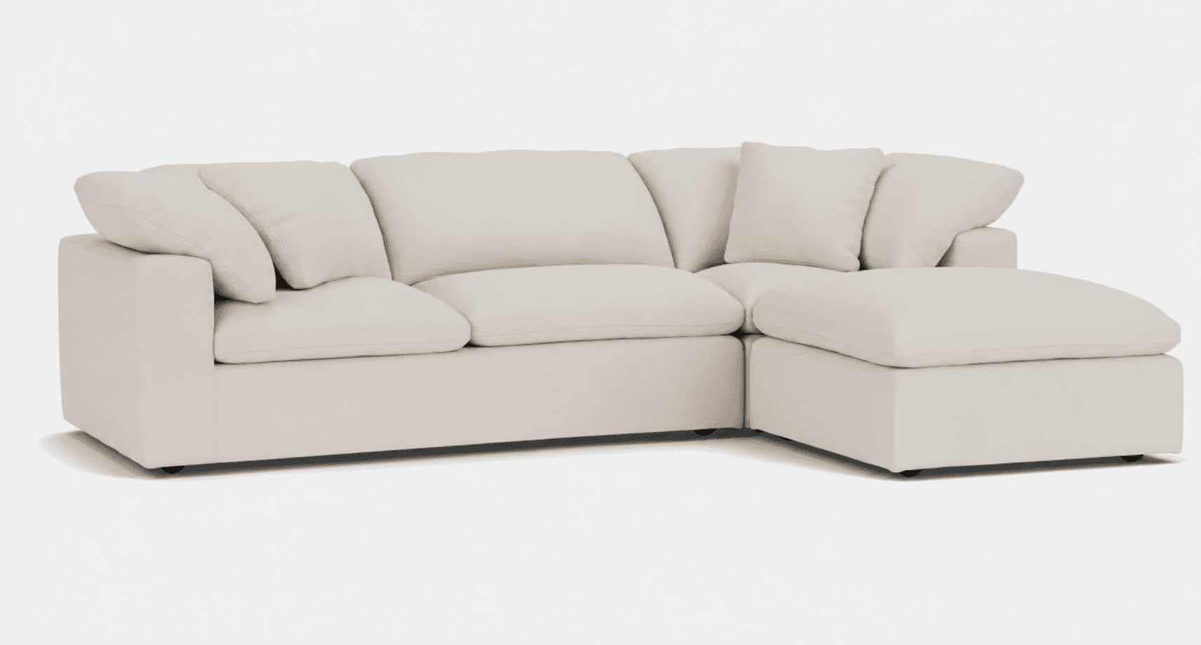 A light-colored, contemporary modular sectional sofa featuring plush, oversized cushions and an integrated chaise lounge on the right side. The sofa exhibits a low-profile design and is upholstered in a soft, cream-colored fabric, likely a linen or cotton blend, offering a relaxed aesthetic. The construction appears to be modular, allowing for flexible arrangement. — 3D model
