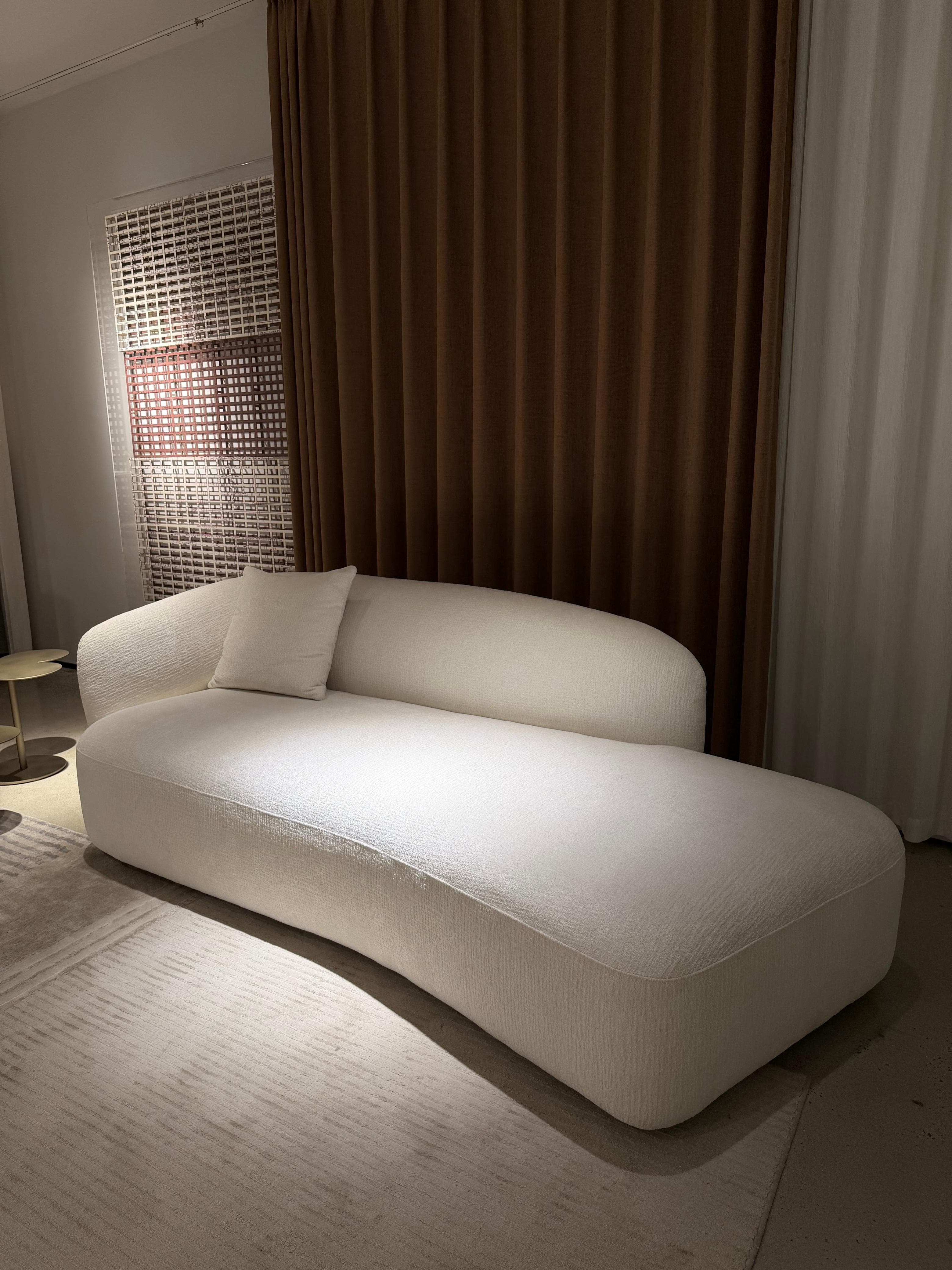 An asymmetrical upholstered chaise lounge in a cream-colored textured fabric, featuring a single integrated armrest/backrest and a curved, elongated form for reclining. — 3D model