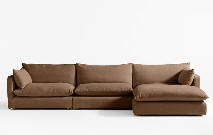 A large, modular sectional sofa upholstered in a neutral taupe-brown fabric. It features a three-seat configuration with a right-hand facing chaise lounge, offering ample seating. The design is characterized by its low-profile frame, deep, plush seat and back cushions, and wide, flat armrests on the left end. The overall aesthetic is contemporary and emphasizes comfort. — 3D model