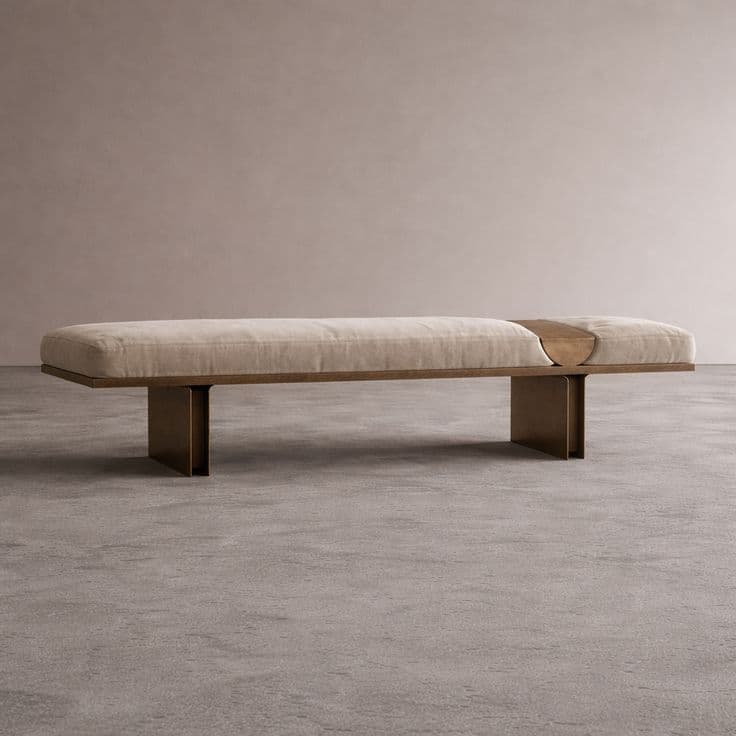 A modern bench with a plush, upholstered seat and a metallic base. The bench features a continuous, elongated cushion in a light neutral tone. The base is constructed from two vertical, rectangular supports connected by a horizontal frame, all appearing to be made of metal with a brushed or patinated finish. A segment of the cushion on the right side is separated by a curved wooden insert, adding a design detail. — 3D model