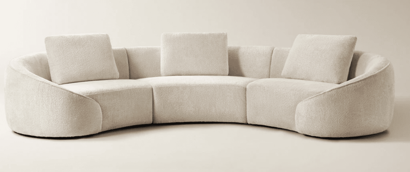 A large, contemporary curved sectional sofa upholstered in a light, textured bouclé fabric, featuring a low profile, integrated armrests, and three loose back cushions. — 3D model