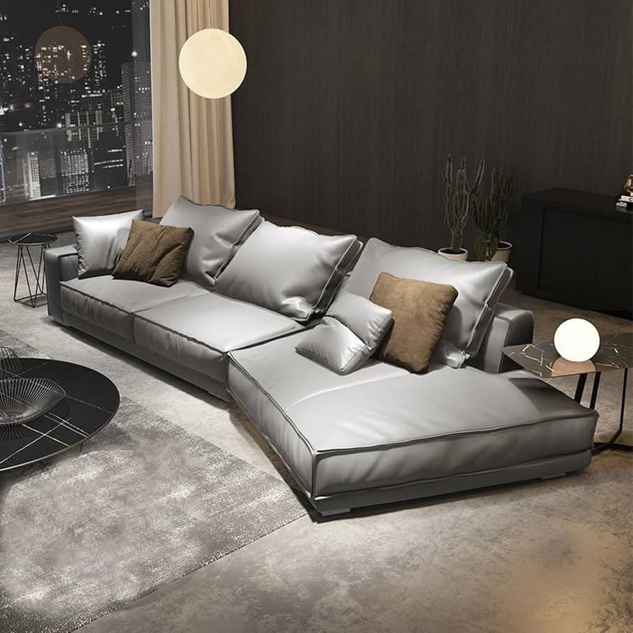 A modern L-shaped sectional sofa upholstered in a light grey leather or high-quality faux leather, featuring plush, soft cushions and contrasting accent pillows. The sofa has a low-profile design with distinct piping along the edges of the cushions and frame. — 3D model