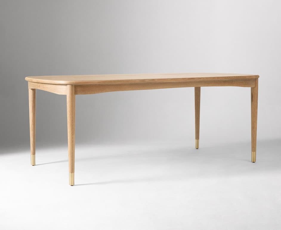 A light-colored wooden dining table with a rectangular top and gently rounded edges. It features four tapered legs, each capped with a brass accent at the base. The legs are positioned slightly inward from the corners of the tabletop. — 3D model