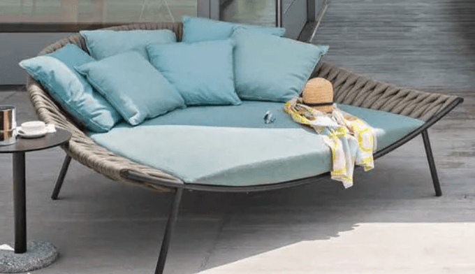 A modern outdoor daybed featuring a semi-circular form with a tightly woven gray synthetic rope backrest and dark metal legs. It is fitted with a large, light blue primary cushion and several smaller matching throw pillows. A straw hat and a patterned towel are placed on the cushion, suggesting a leisure context. — 3D model