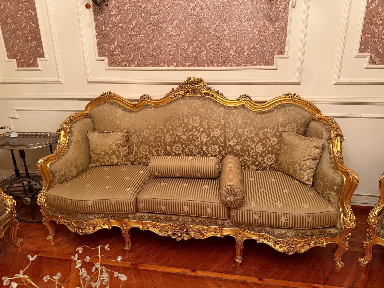 An opulent three-seater sofa characterized by an elaborate, hand-carved wooden frame finished in gold leaf. The sofa features a curvilinear backrest, armrests, and apron adorned with intricate scrollwork. It is upholstered in a rich, patterned beige and gold fabric, with striped seat cushions. The piece rests on decorative cabriole legs and includes two square throw pillows and two cylindrical bolster pillows. — 3D model