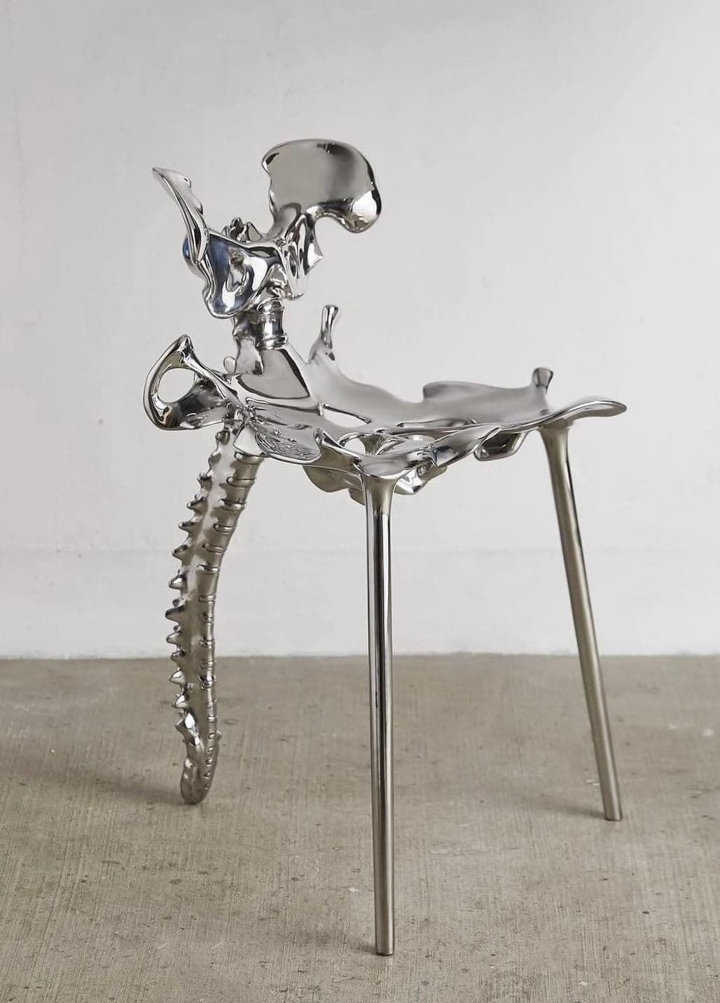 A unique chair with a design inspired by skeletal anatomy. The seat and backrest appear to be sculpted in the form of pelvic bones and a spine, while the legs are slender and metallic. The overall finish is highly reflective, giving it a polished metallic appearance. — 3D model