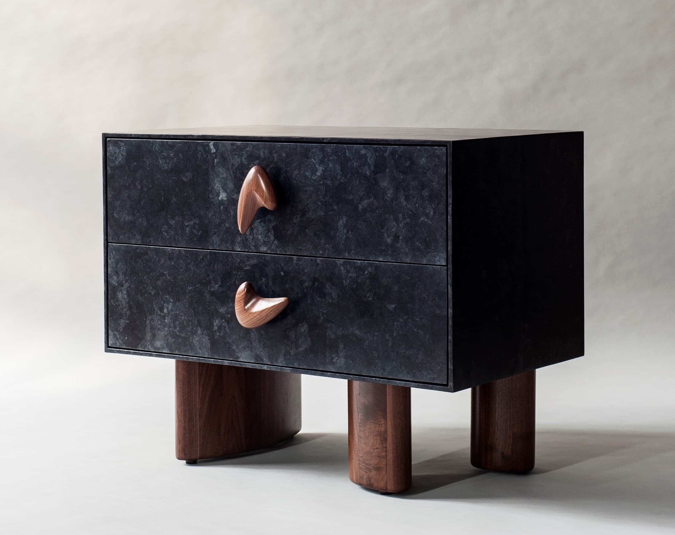 A contemporary nightstand featuring a dark, textured exterior with two drawers. Each drawer is adorned with a unique, sculpted wooden handle. The nightstand is supported by four distinct wooden legs, two of which are thicker and curved, while the front two are cylindrical. The overall design suggests a modern aesthetic with organic influences. — 3D model